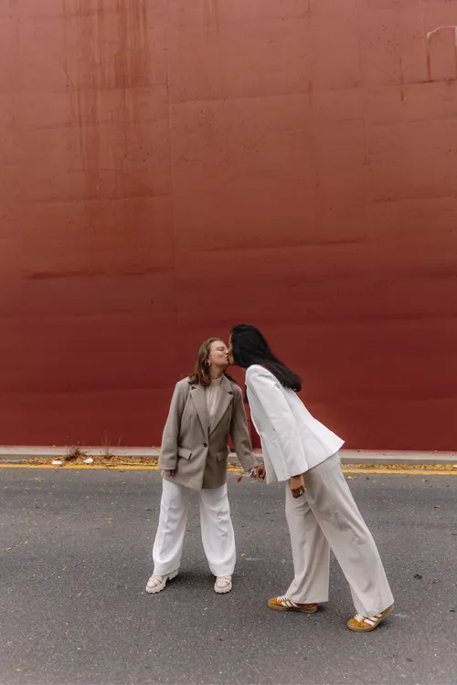 Bruna e Clara se beijando de mãos dadas diante do muro terracota